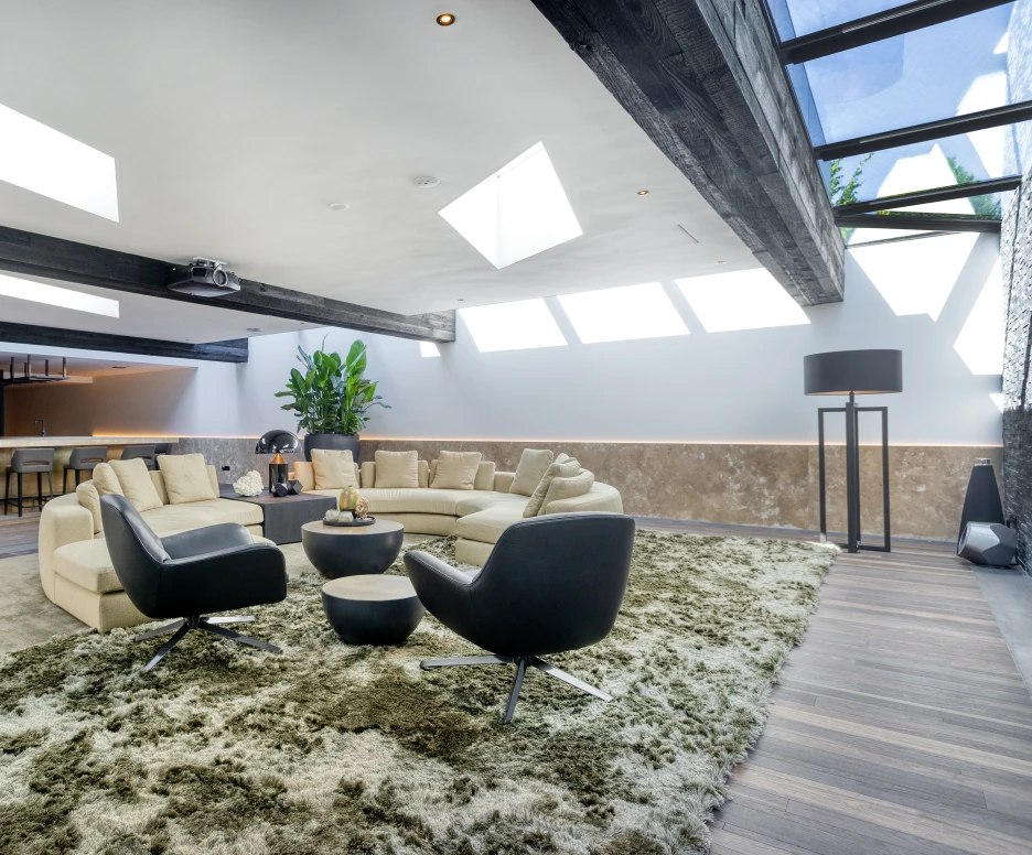 A modern living room featuring a circular beige sofa and two black swivel chairs around a round coffee table. The room has large skylights, a plush green rug, and a bar area with pendant lights in the background. Lush greenery adds a touch of nature to the space.