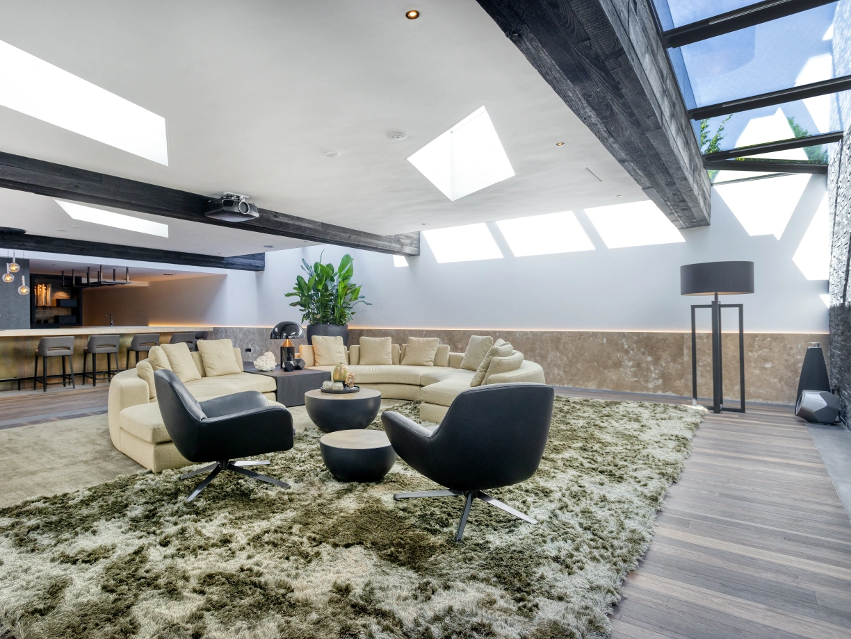 Salon moderne avec un canapé en forme de L, deux fauteuils noirs et un grand tapis moelleux. La pièce est lumineuse grâce aux lucarnes et présente des touches végétales ainsi qu’un bar en arrière-plan.