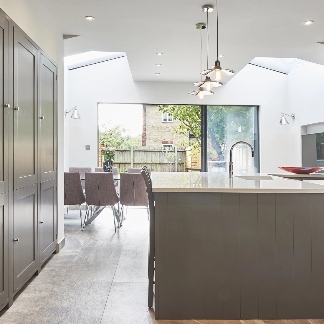 Modern kitchen with gray cabinetry, an island with a countertop, and a view of a dining area through large glass doors. The space is well-lit with pendant lighting and features a mix of natural and artificial light.