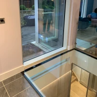 Large modern window with a view of a patio and garden. A reflection of a person is visible on the glass. The interior features a sleek design with a glass floor and grey tiles.