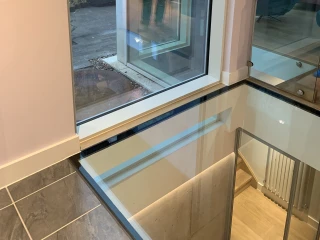 Ventana grande con un marco moderno, mostrando el exterior de una casa y el reflejo de una persona. El área interior cuenta con un piso de baldosas y una escalera de vidrio.