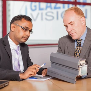 Two men in business attire are engaged in a discussion at a conference table. One man is pointing to a document while the other examines a gray project sample. A "Glazing Vision" sign is visible in the background.