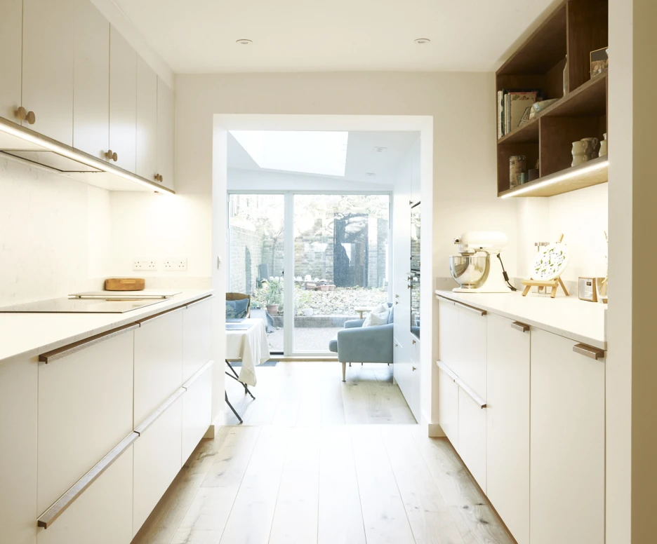 A modern kitchen with white cabinetry and a light wood floor. The view opens up to a bright living space with a garden visible through large glass doors. Natural light fills the area, highlighting a countertop and decorative items on shelves.