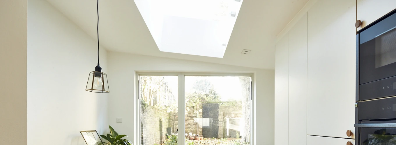 A bright, modern kitchen features a skylight, wooden flooring, and white cabinetry. A small table with a plant is beside a cozy blue armchair, while sliding glass doors open to a garden space filled with greenery.