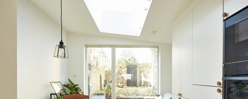 A bright, modern kitchen features a skylight, wooden flooring, and white cabinetry. A small table with a plant is beside a cozy blue armchair, while sliding glass doors open to a garden space filled with greenery.