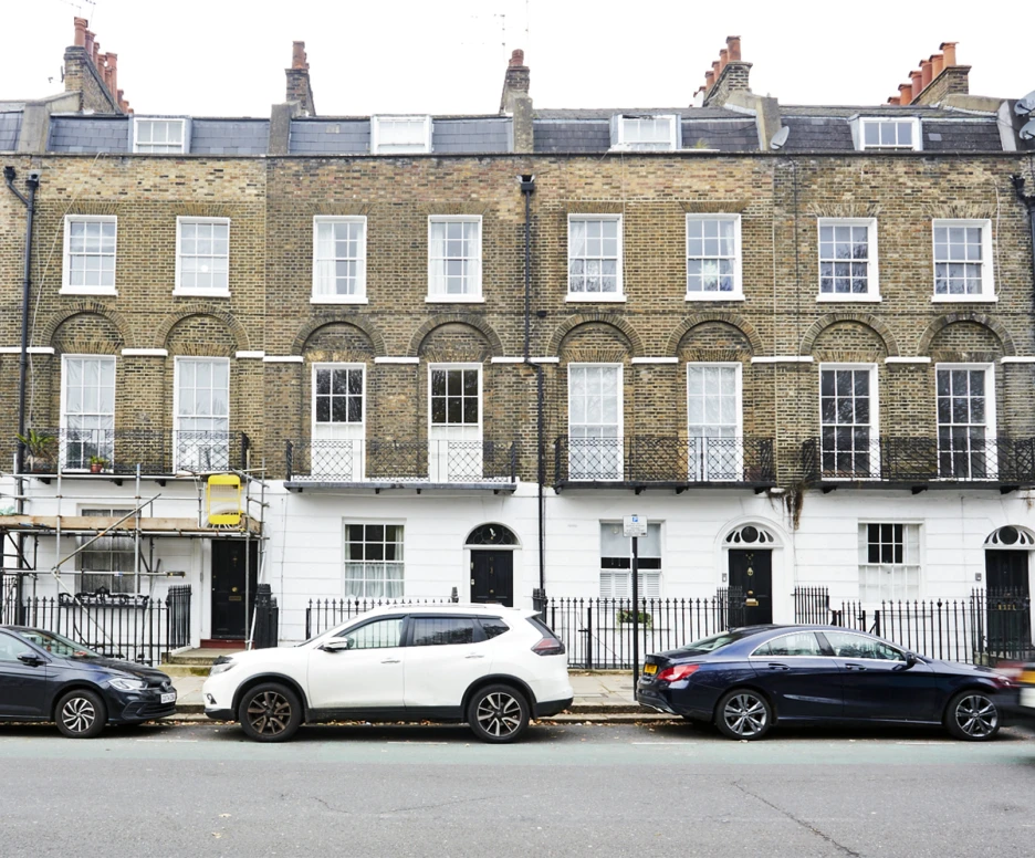 A row of traditional brownstone houses with white trim, featuring multiple windows and several doors. Parked cars line the street in front, with scaffolding visible on one of the buildings.