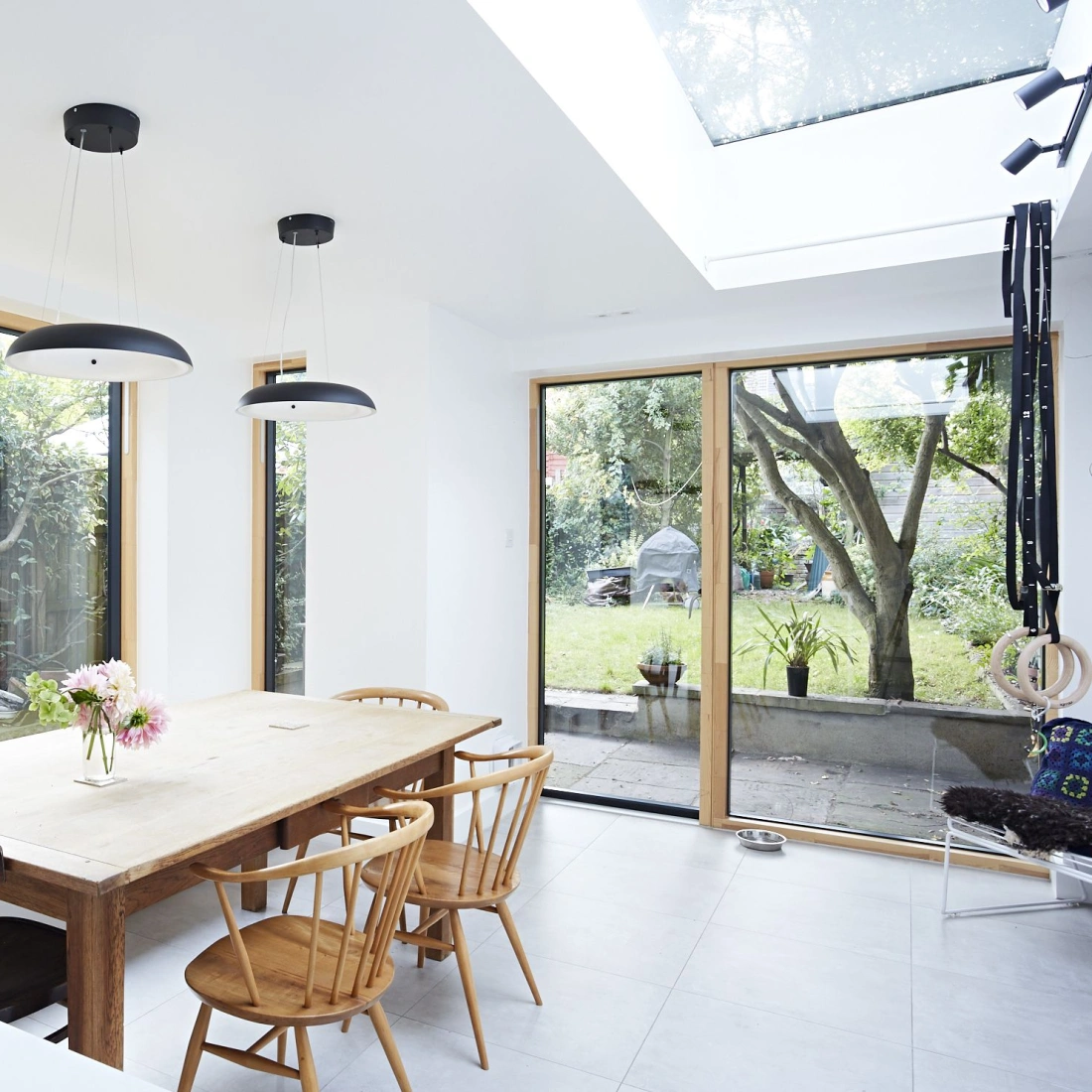 A bright dining area featuring a wooden table surrounded by chairs. Large windows offer a view of a lush garden and allow natural light to flood the space. Two modern pendant lights hang above the table.