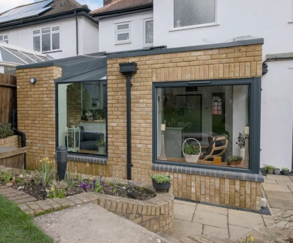 A modern brick extension on a house features large glass windows that provide a view into a cozy interior. The outdoor area includes a pathway, garden beds with flowers, and green grass. Light clouds are visible in the blue sky above.