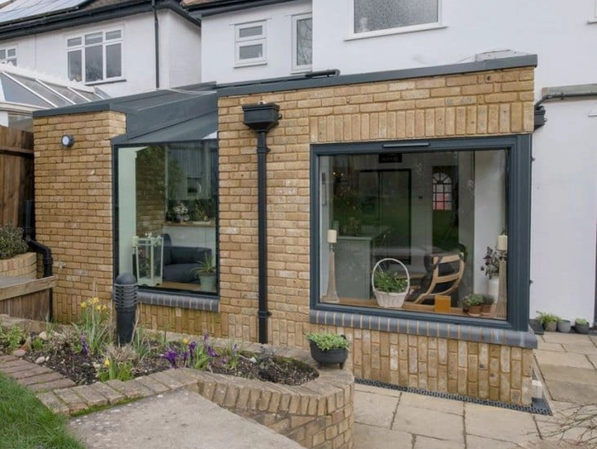 A modern brick extension on a house features large glass windows that provide a view into a cozy interior. The outdoor area includes a pathway, garden beds with flowers, and green grass. Light clouds are visible in the blue sky above.