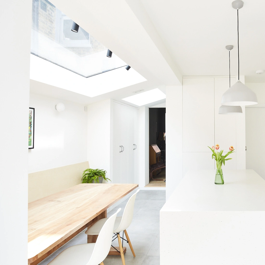 A bright, modern kitchen featuring a large white island, sleek cabinetry, and a dining table made of wood. A skylight illuminates the space, and a vase of tulips adds a touch of color. The walls are painted white, with a pink accent behind the stove.