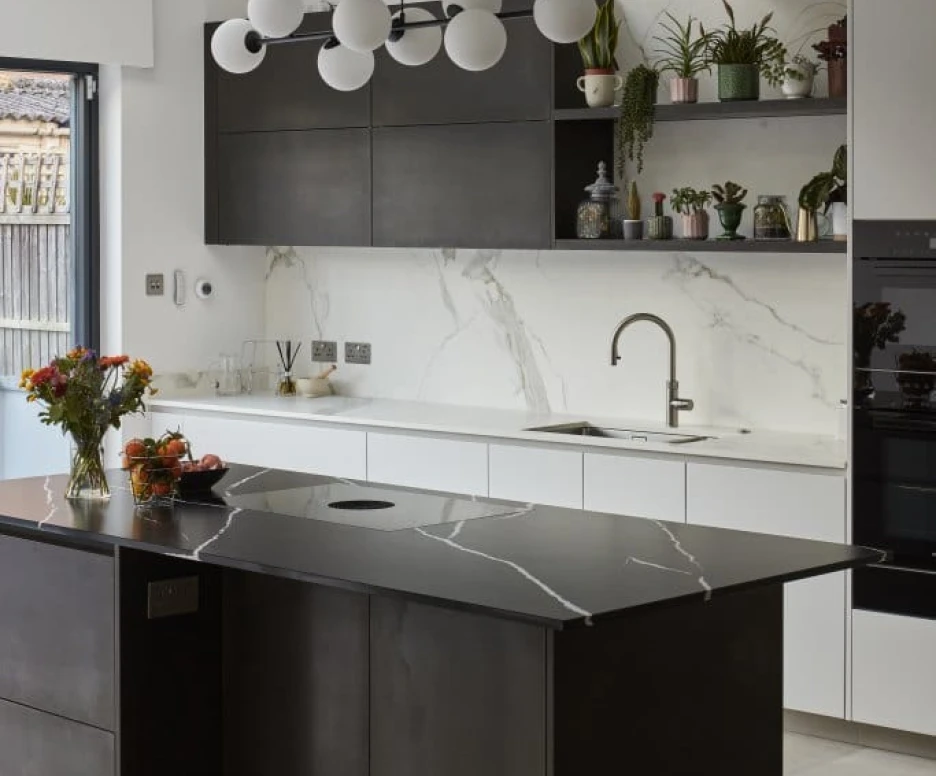 A modern kitchen featuring a sleek black island with a unique marble countertop. Above, a contemporary chandelier with white globes hangs. The walls are adorned with marble and dark cabinetry, while shelves display potted plants and decorative items. Fresh flowers and fruits are arranged on the island.