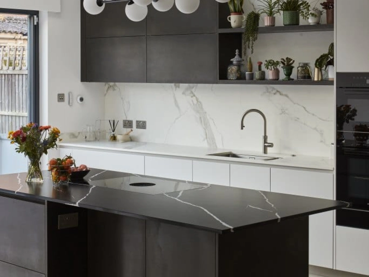 A modern kitchen featuring a sleek black island with a unique marble countertop. Above, a contemporary chandelier with white globes hangs. The walls are adorned with marble and dark cabinetry, while shelves display potted plants and decorative items. Fresh flowers and fruits are arranged on the island.
