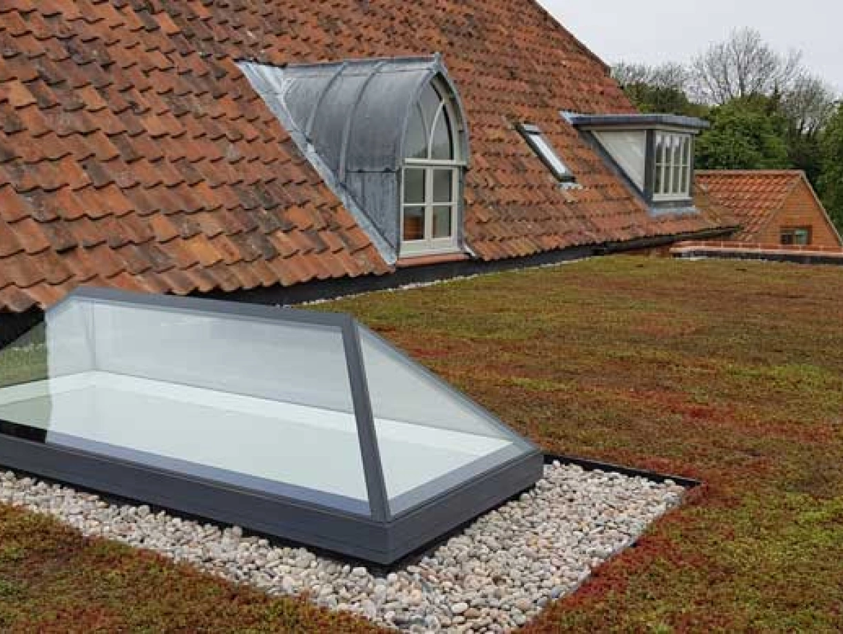 A green flat roof with a skylight, surrounded by small stones. The roof features terracotta tiles and a sloped section with dormer windows.