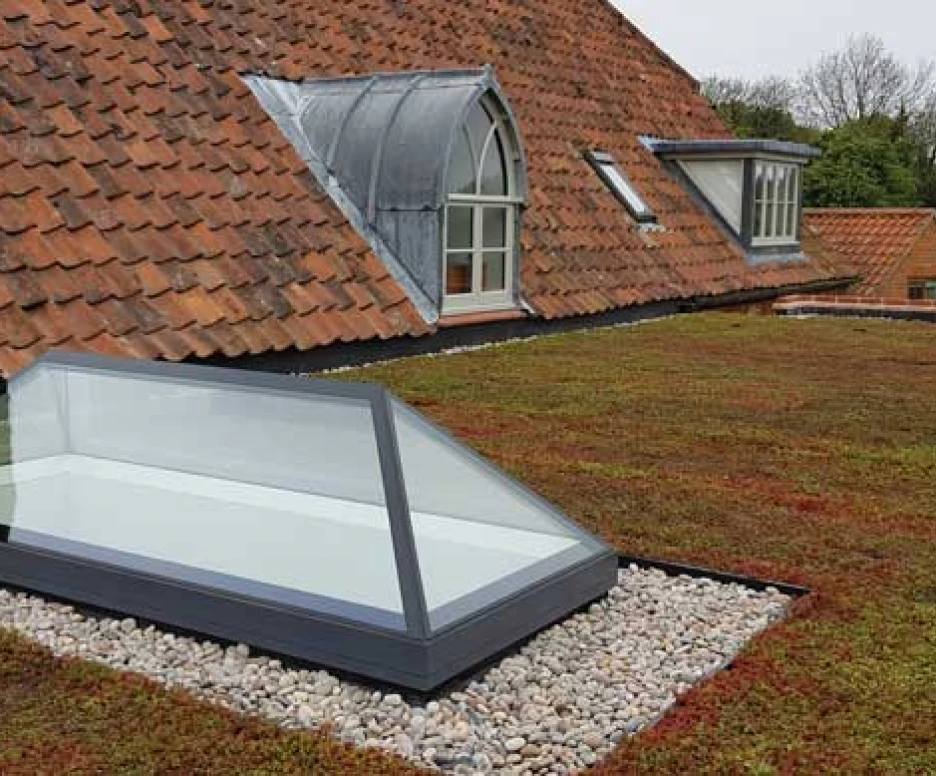 A green flat roof with a skylight, surrounded by small stones. The roof features terracotta tiles and a sloped section with dormer windows.