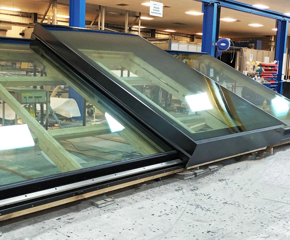 Three large slanted glass panels are arranged together on a wooden frame in a workshop. The panels are partially illuminated by overhead lights, showcasing their sleek edges and reflective surfaces. Tools and materials are visible in the background, indicating an active construction or assembly area.