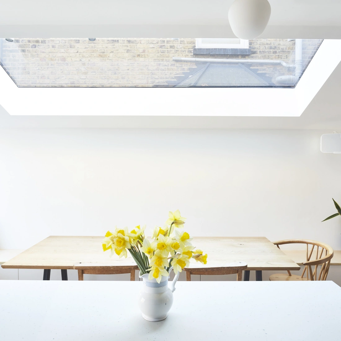 Bright, minimalist kitchen featuring a large skylight, wooden dining table with chairs, and a vase of yellow flowers on the counter. A potted plant adds a touch of greenery.