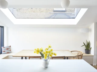 Bright kitchen featuring a large skylight above a wooden dining table with chairs. A vase of yellow daffodils sits on a countertop, and there's a potted plant in the corner. A patterned cushion is on a nearby bench.
