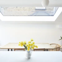 Bright, minimalist kitchen featuring a large skylight, wooden dining table with chairs, and a vase of yellow flowers on the counter. A potted plant adds a touch of greenery.