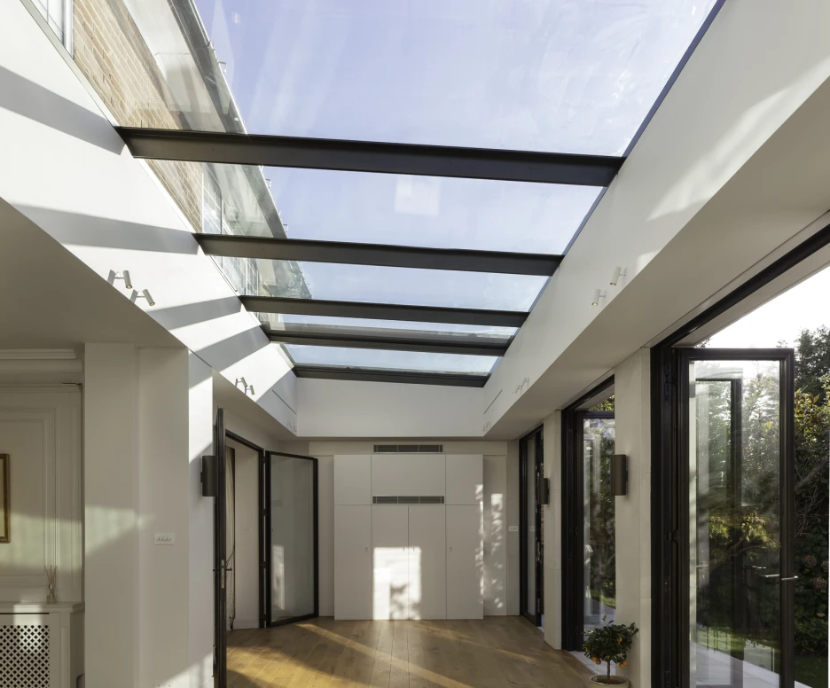 Bright interior space with a glass ceiling and large windows. Sunlight casts shadows on the wooden floor, creating a warm atmosphere. Modern design includes open doors leading to an outdoor area.