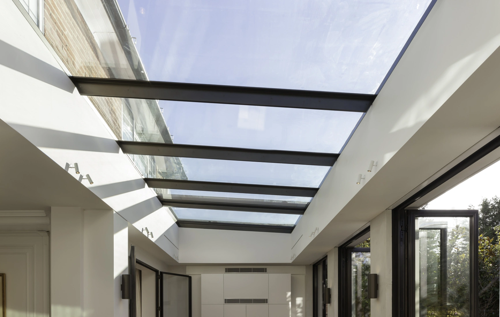 Interior de una habitación con gran entrada de luz natural a través de un techo de cristal. Suelo de madera clara y puertas de vidrio que se abren hacia el exterior. La decoración es minimalista con un toque moderno.