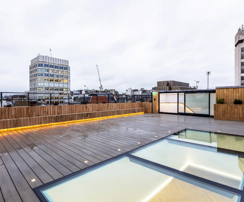 A modern rooftop terrace featuring wooden decking and glass panels, with a city skyline visible in the background. Overcast skies and crane silhouettes add to the urban atmosphere.