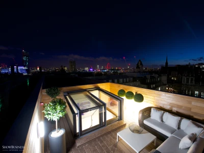 A stylish rooftop terrace with modern furniture, featuring a cozy seating area and potted plants. The skyline of a city is visible in the background, illuminated against a twilight sky, showcasing iconic structures and a ferris wheel.