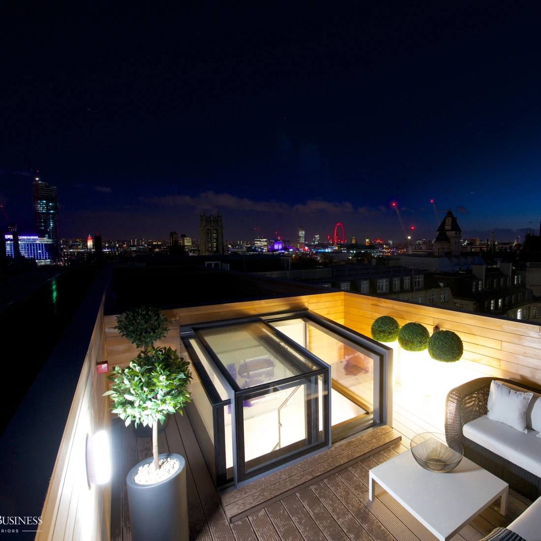 A stylish rooftop terrace with modern furniture, featuring a cozy seating area and potted plants. The skyline of a city is visible in the background, illuminated against a twilight sky, showcasing iconic structures and a ferris wheel.