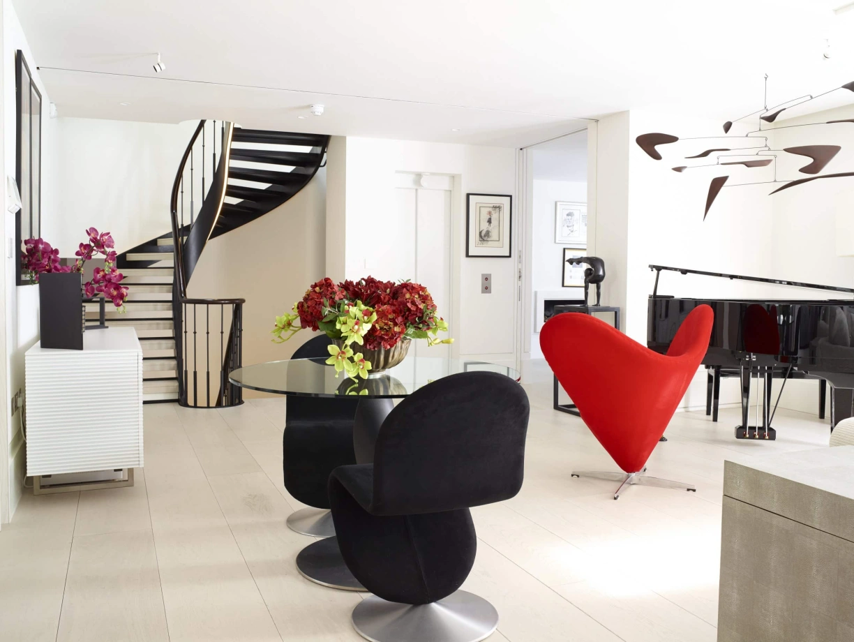 A modern living space with a distinctive heart-shaped red chair, a round glass dining table surrounded by black chairs, and vibrant flowers in vases. A spiral staircase and a grand piano are visible in the background, adding elegance to the bright, open layout.