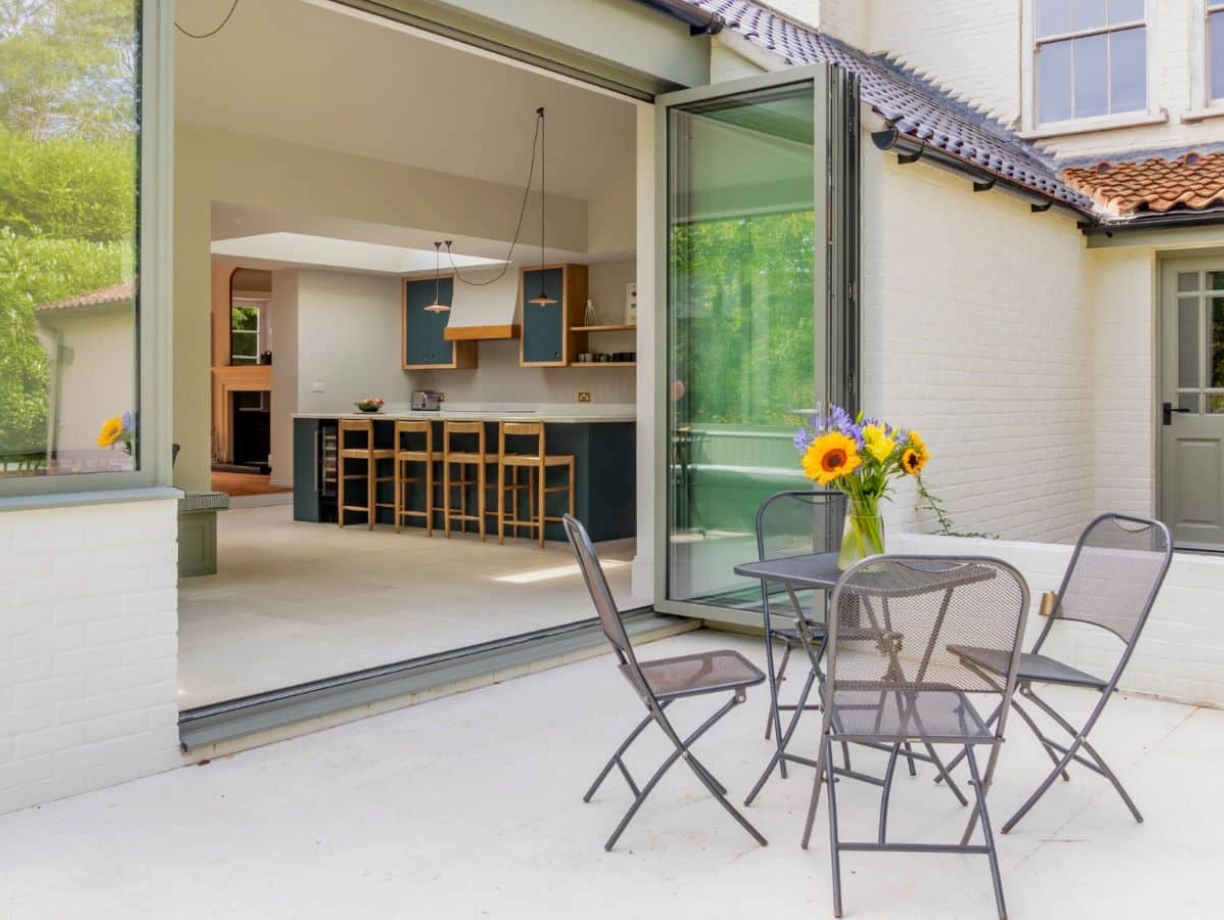 A cozy outdoor patio with a table and four metal chairs. A vase of sunflowers sits on the table. Open glass doors lead into a modern kitchen area with wooden bar stools and a bright interior. Lush greenery can be seen outside.