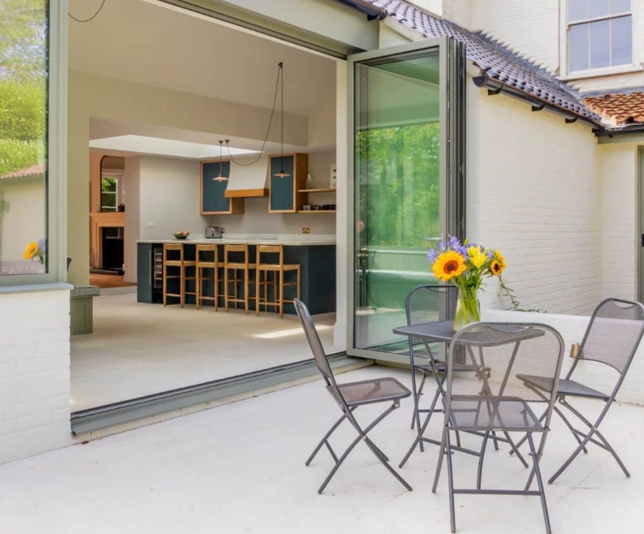 A cozy outdoor patio with a table and four metal chairs. A vase of sunflowers sits on the table. Open glass doors lead into a modern kitchen area with wooden bar stools and a bright interior. Lush greenery can be seen outside.