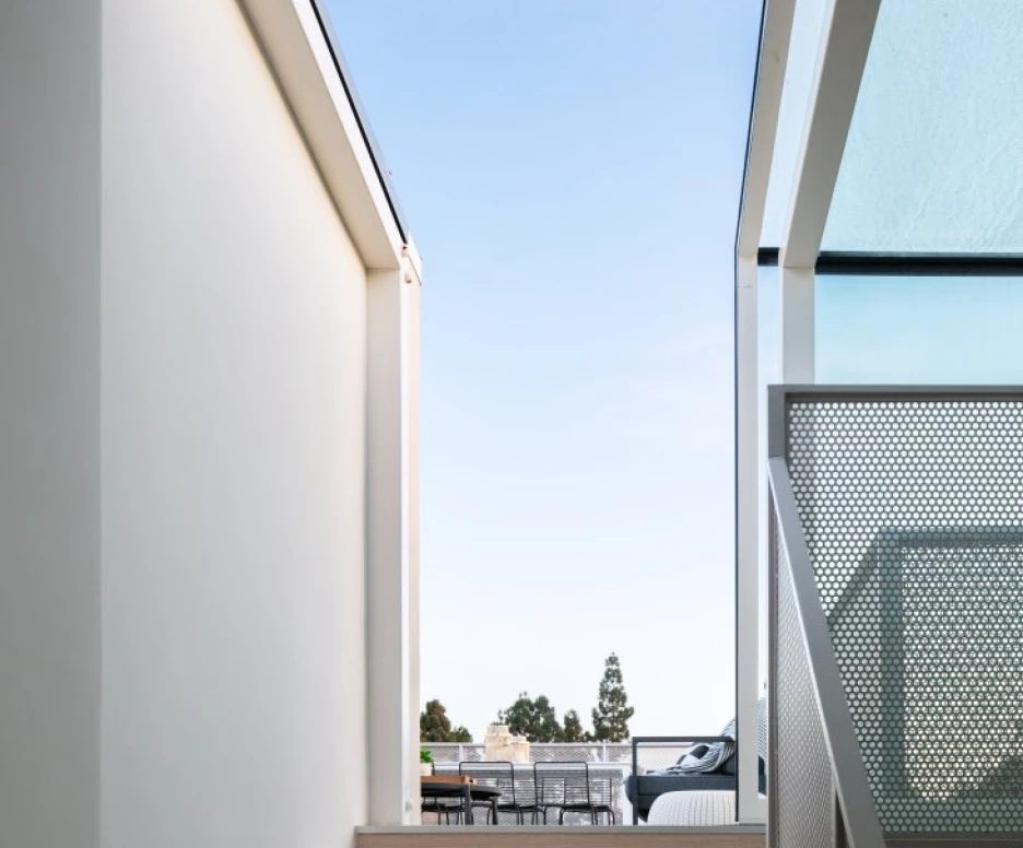 A staircase leads up from a narrow corridor, framed by two white walls, towards a clear blue sky. A modern balcony with outdoor seating is visible in the background.