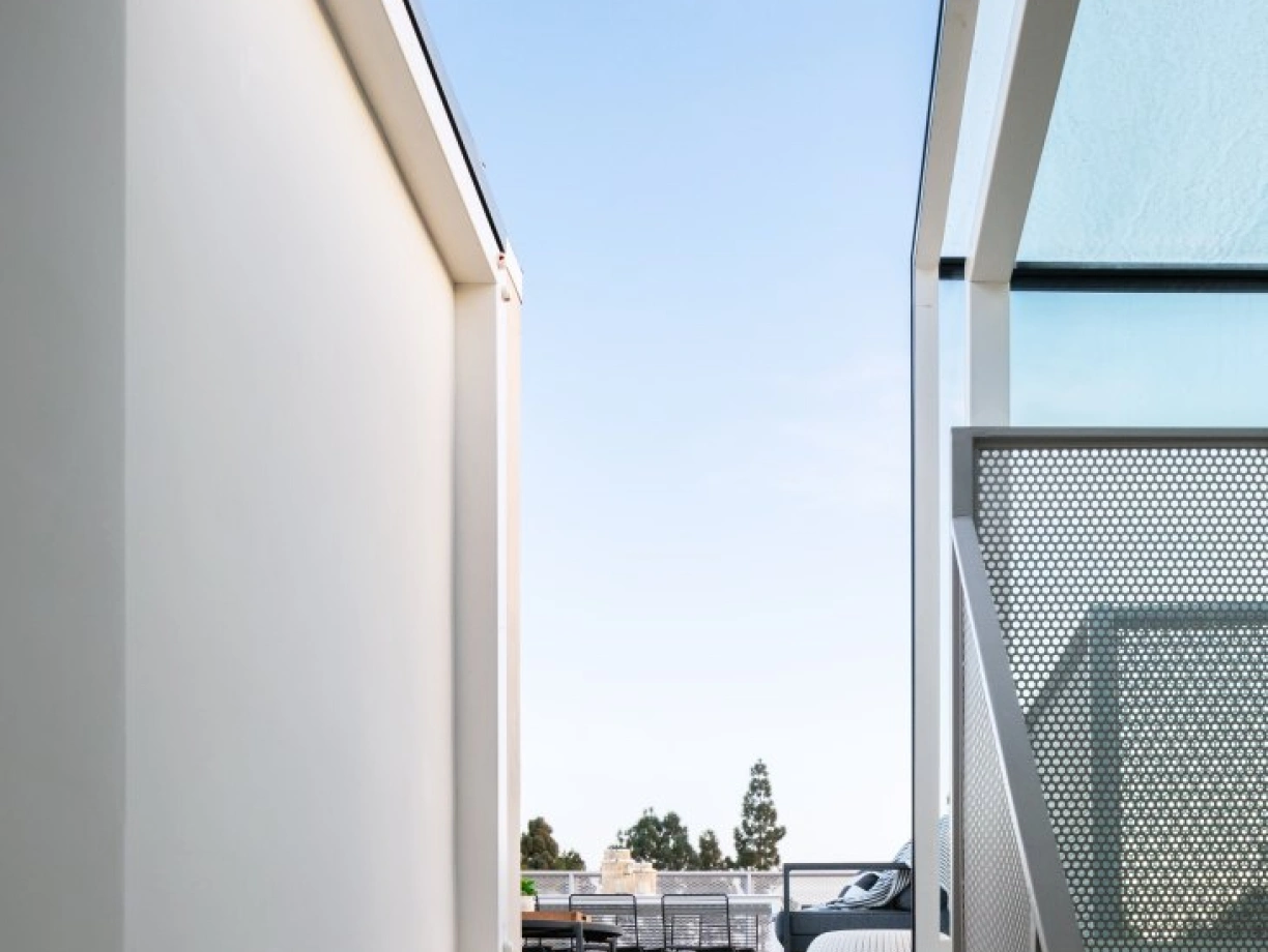 A staircase leads up from a narrow corridor, framed by two white walls, towards a clear blue sky. A modern balcony with outdoor seating is visible in the background.