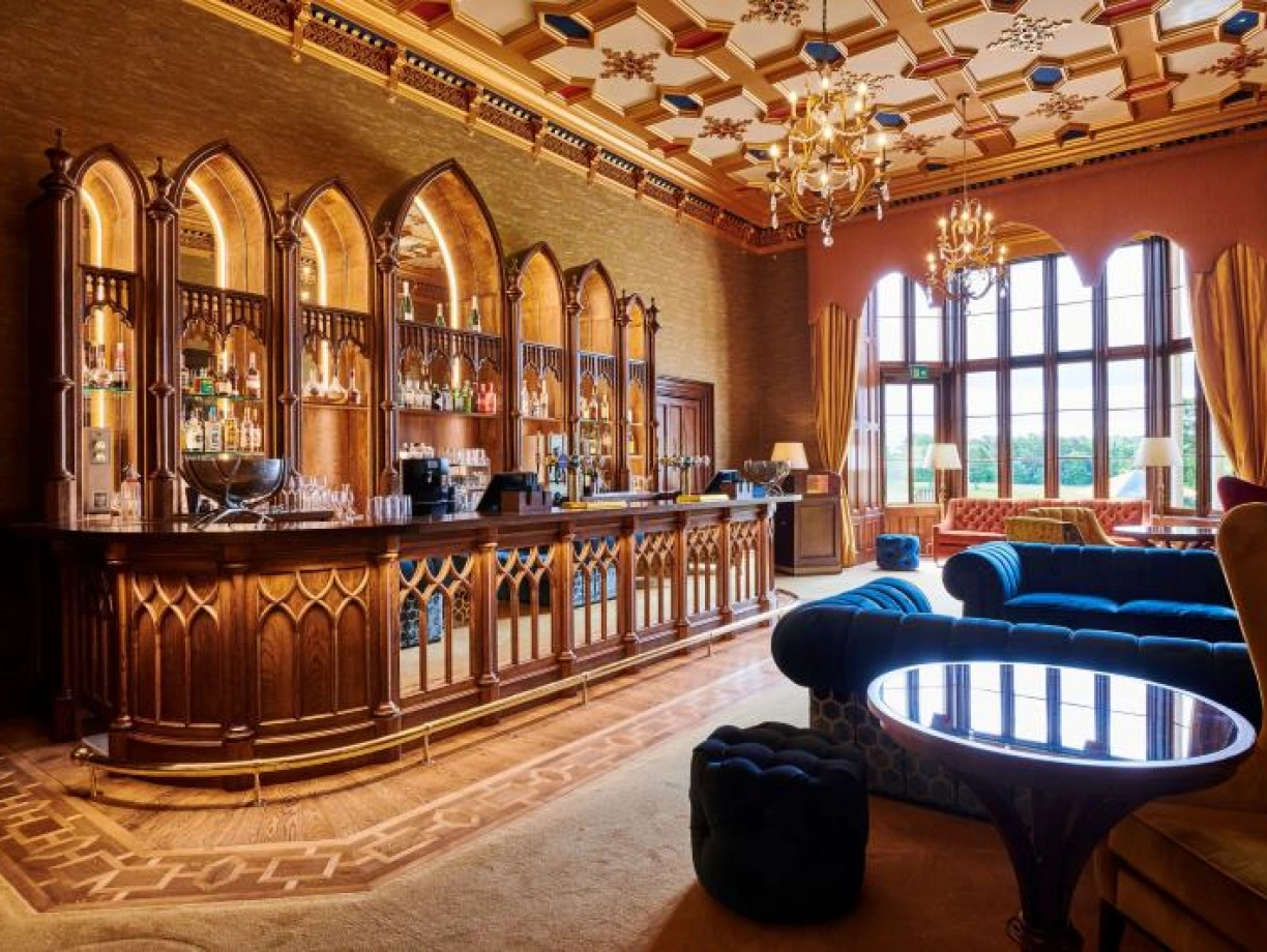 A luxurious hotel bar featuring intricate woodwork, arched windows, and a variety of bottles displayed behind the counter. Plush seating in rich colors complements the elegant decor, with a chandelier hanging overhead.