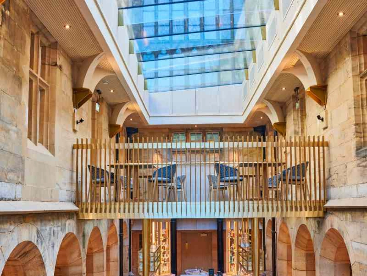 Restaurant élégant avec une structure en pierre et de grandes fenêtres. La lumière naturelle illumine l’espace, mettant en valeur un bar central et des tables disposées en bas. Un balcon en bois se trouve à l’étage supérieur, ajoutant une touche moderne à l’architecture.