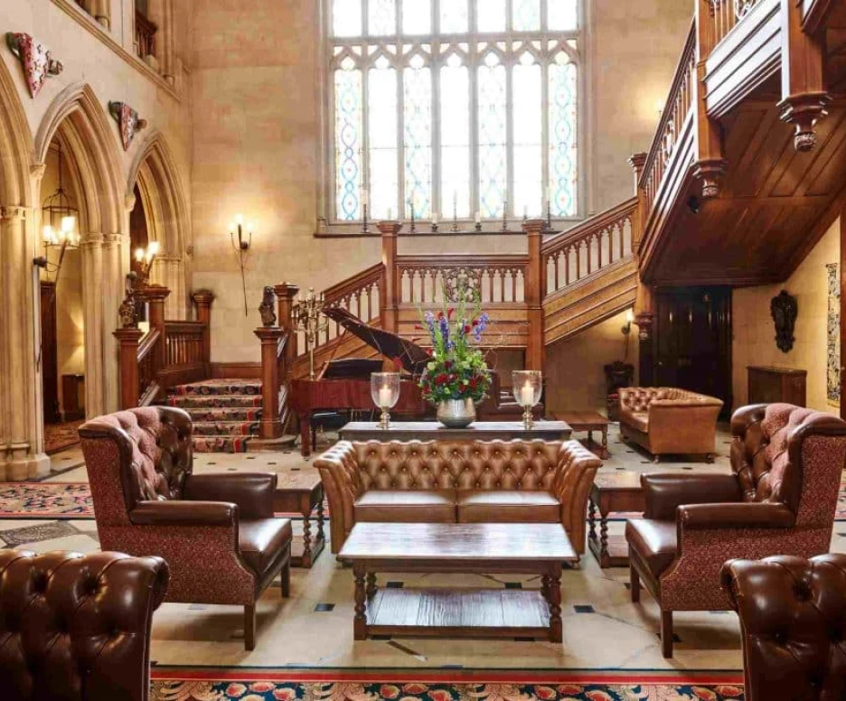 Elegant lobby featuring ornate wooden stairs, a grand piano, and plush leather armchairs. A large stained glass window adds natural light, and colorful floral arrangements decorate the coffee table.