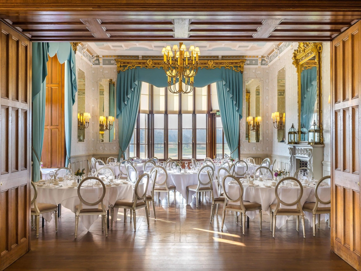 An elegantly decorated dining room featuring round tables set with white tablecloths, fine china, and glassware. Large windows with turquoise drapes allow natural light to illuminate the space. The walls are adorned with ornate moldings and a fireplace, enhancing the room's sophistication.