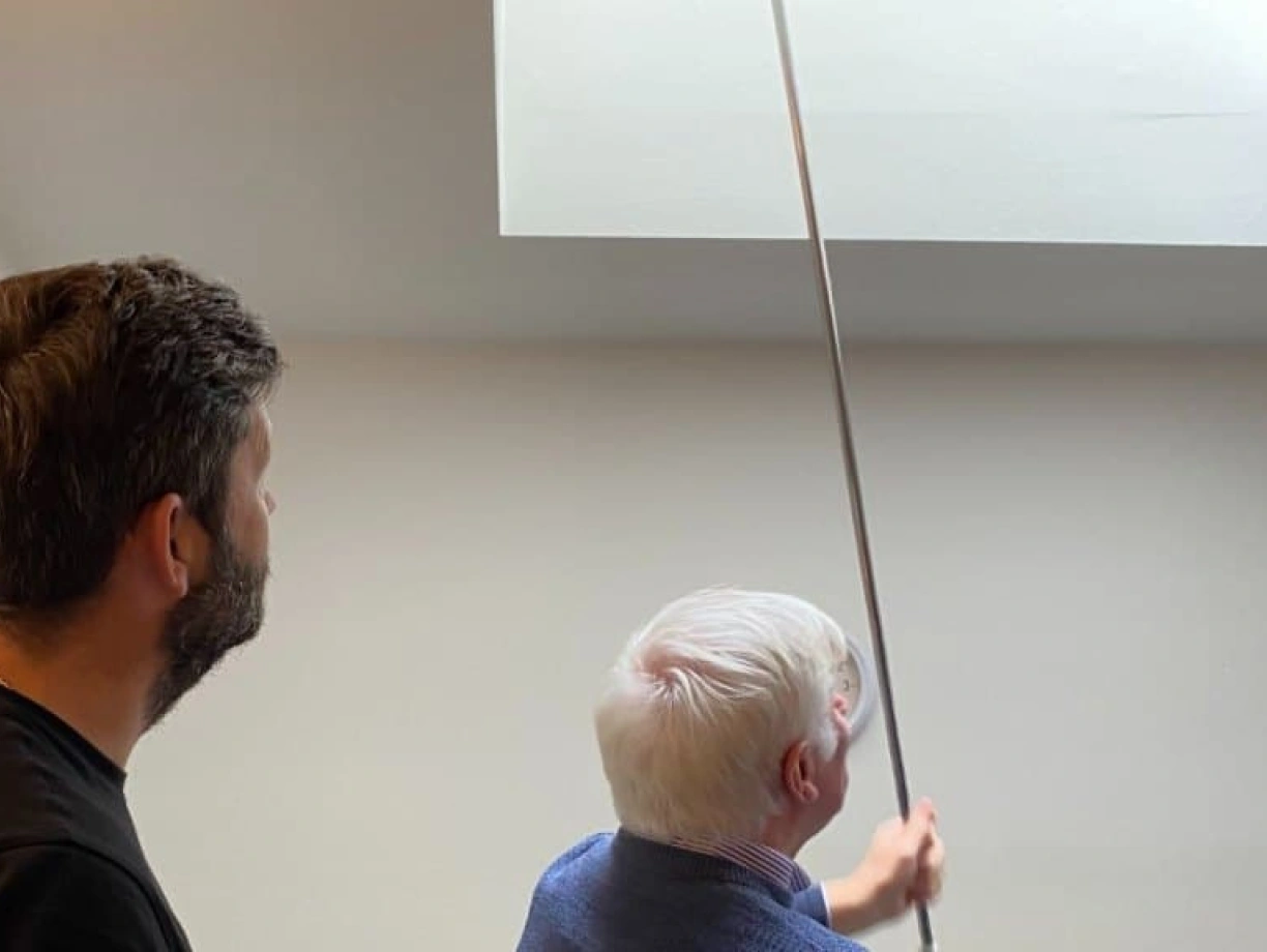 A younger man and an older man are looking up at a ceiling while the older man extends a pole towards a high corner. The room has light-colored walls and appears to be indoors.