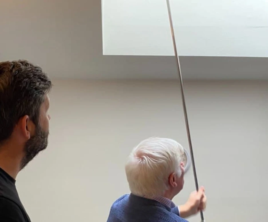 A younger man and an older man are looking up at a ceiling while the older man extends a pole towards a high corner. The room has light-colored walls and appears to be indoors.