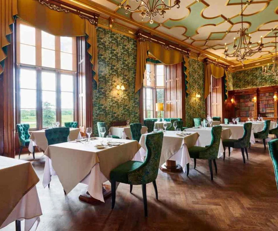 Salle de restaurant élégante avec des tables dressées, des chaises vertes en velours et des murs tapissés de motifs floraux. De grandes fenêtres laissent entrer la lumière naturelle, offrant une vue sur l’extérieur.