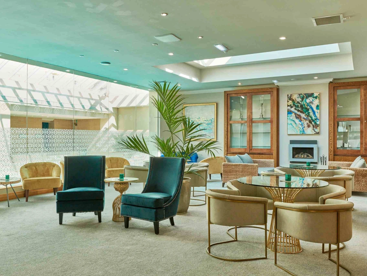 A modern lounge with a mix of plush seating options, including blue and beige chairs and circular tables. Large windows allow natural light to fill the space, highlighting a contemporary artwork on the wall and a decorative plant in the corner.