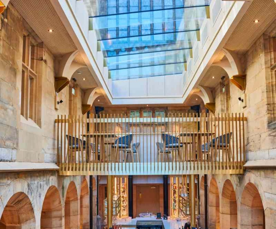 A spacious restaurant interior featuring a high glass ceiling allowing natural light to flood in. The design includes stone arches and a central bar area surrounded by tables set elegantly. Wooden railings and modern furniture add a contemporary touch to the historic architecture.