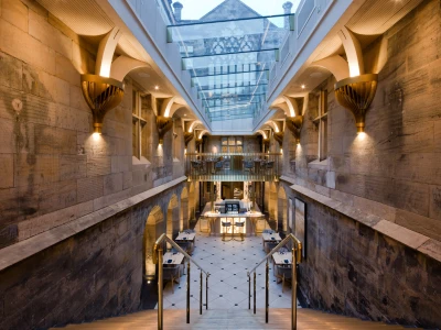 A spacious interior featuring stone walls and elegant architectural details. A grand staircase leads down to a dining area with modern tables and chairs, illuminated by warm lighting. Above, a glass ceiling allows natural light to enter the space.