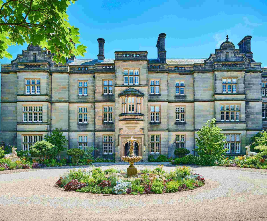 Historic stone building with intricate architecture, surrounded by colorful gardens and trees. A central fountain adds charm to the landscaped area in front. Bright blue sky enhances the scene.