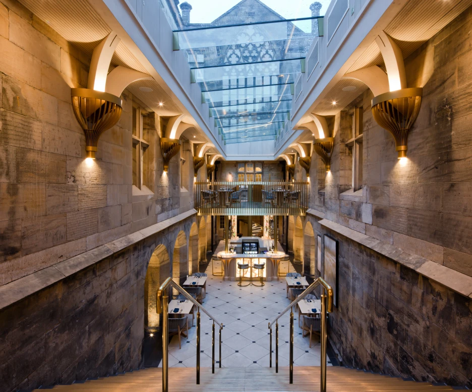 A spacious interior featuring stone walls and elegant architectural details. A grand staircase leads down to a dining area with modern tables and chairs, illuminated by warm lighting. Above, a glass ceiling allows natural light to enter the space.