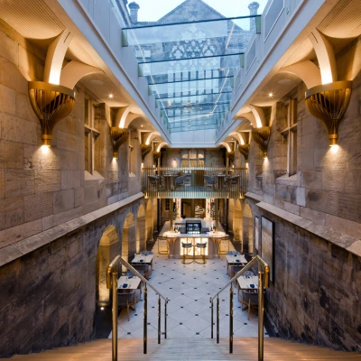 A spacious interior with a staircase leading down to a lively dining area. The walls feature exposed stone, and large windows allow natural light to fill the space. Elegant lighting and modern furnishings complement the historic architecture.