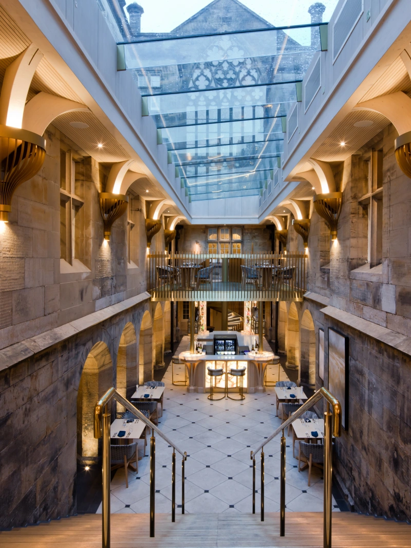 A spacious interior featuring stone walls and elegant architectural details. A grand staircase leads down to a dining area with modern tables and chairs, illuminated by warm lighting. Above, a glass ceiling allows natural light to enter the space.