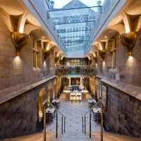 A spacious interior featuring stone walls and elegant architectural details. A grand staircase leads down to a dining area with modern tables and chairs, illuminated by warm lighting. Above, a glass ceiling allows natural light to enter the space.