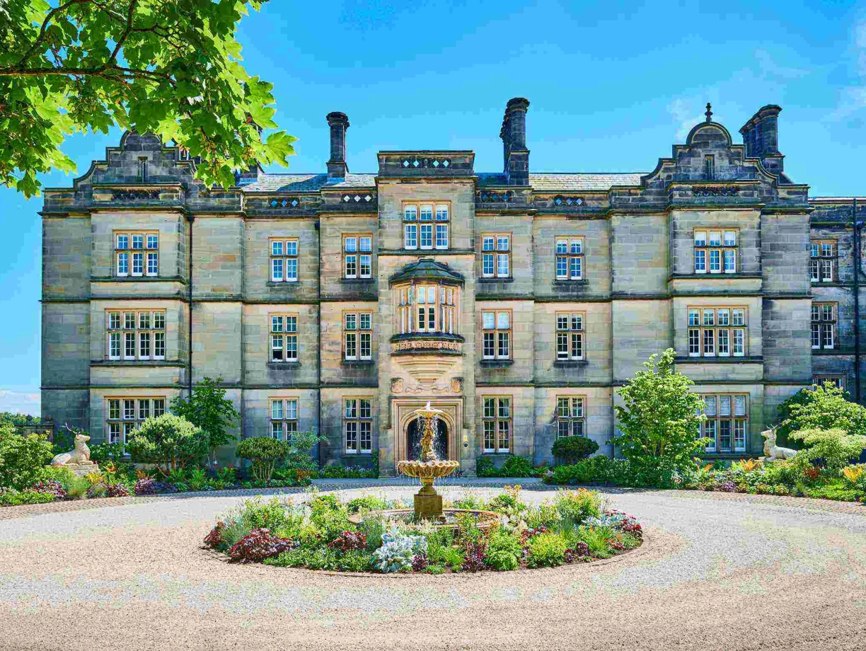 A grand stone building with multiple windows is set against a bright blue sky, surrounded by a circular garden featuring blooming flowers and a central fountain. Lush trees frame the scene, enhancing the picturesque setting.