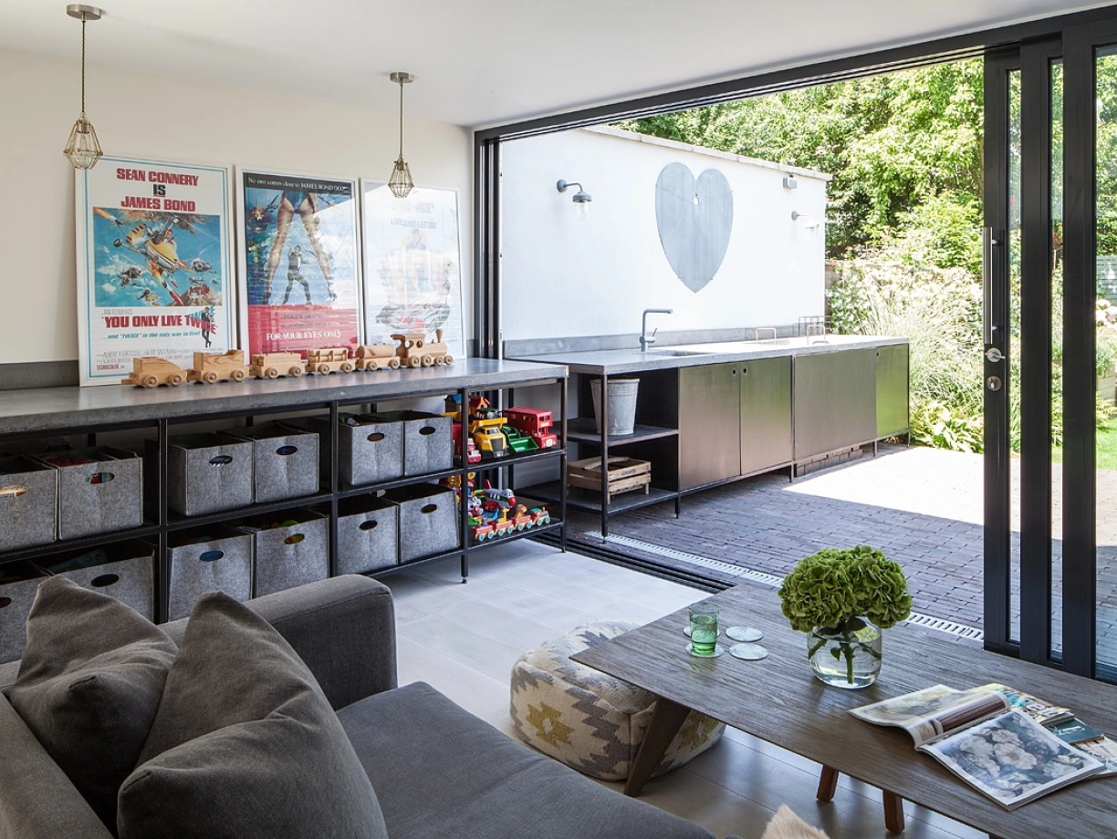 A modern living space with a gray couch and a wooden coffee table. There are storage boxes filled with toys and books. Large sliding glass doors open to an outdoor area featuring a heart-shaped mural on the wall and a sleek outdoor kitchen. Bright natural light floods the room.