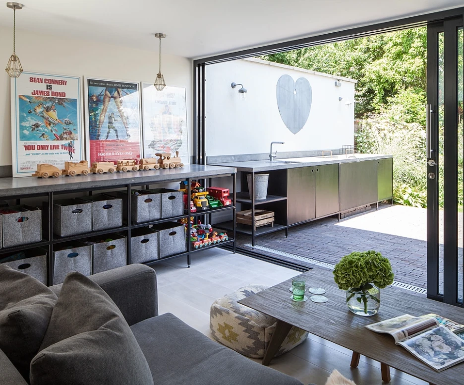 A modern living space with a gray couch and a wooden coffee table. There are storage boxes filled with toys and books. Large sliding glass doors open to an outdoor area featuring a heart-shaped mural on the wall and a sleek outdoor kitchen. Bright natural light floods the room.